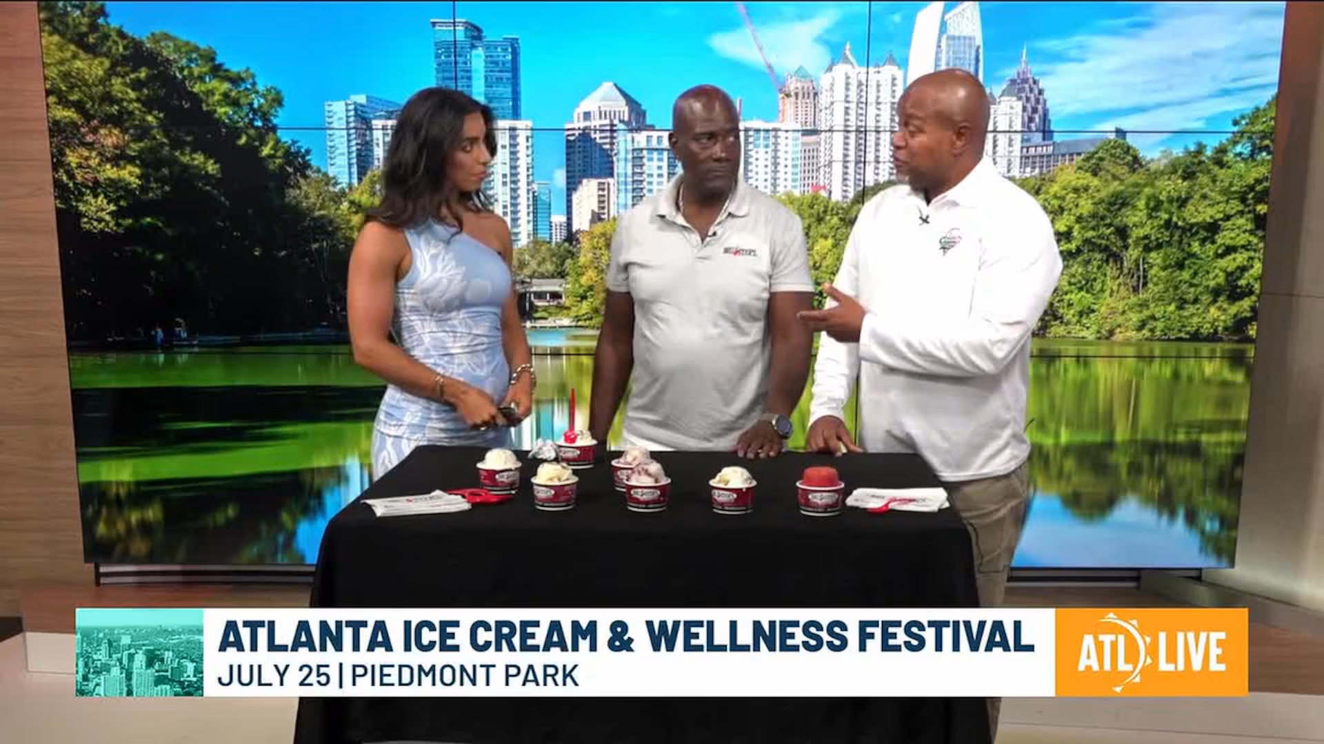 Three people stand behind a table with ice cream cups, discussing the Atlanta Ice Cream & Wellness Festival. A city skyline and park are visible in the backdrop. The atmosphere is lively and engaging.